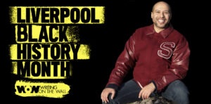 Liverpool Black History Month. Image of Dean Atta, seated wearing a red jacket.
