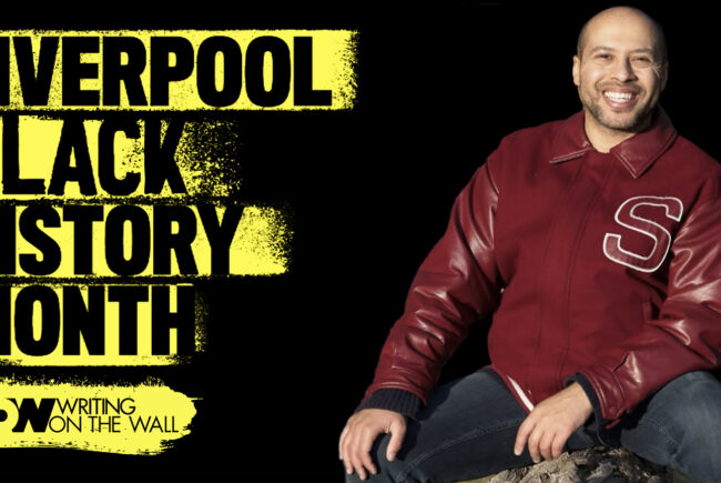 Liverpool Black History Month. Image of Dean Atta, seated wearing a red jacket.